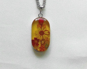 May include: A silver chain necklace with a pendant. The pendant is a clear, oval-shaped resin encasing dried yellow and red flowers. The flowers are visible within the transparent resin, creating a unique floral design.
