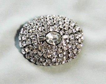 May include: An oval-shaped brooch covered in numerous small, clear, faceted rhinestones. A single, larger, oval-cut clear stone is set prominently in the center, flanked by two small, rectangular, baguette-cut clear stones on either side.