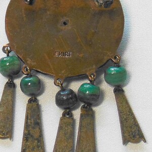 May include: Vintage brass-toned pendant with green and black beads and dangling trapezoid-shaped accents. The pendant is stamped with the word "MEXICO". The design features a circular base with dangling elements.