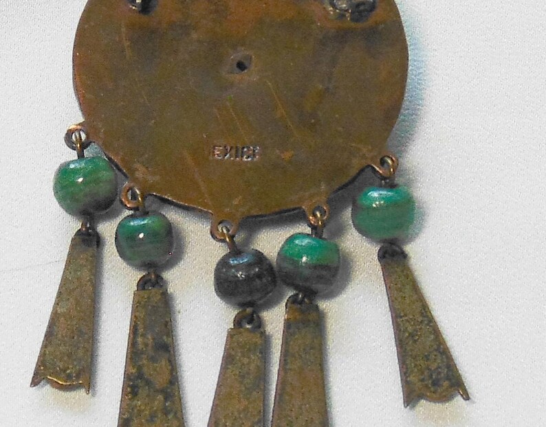 May include: Vintage brass-toned pendant with green and black beads and dangling trapezoid-shaped accents. The pendant is stamped with the word "MEXICO". The design features a circular base with dangling elements.