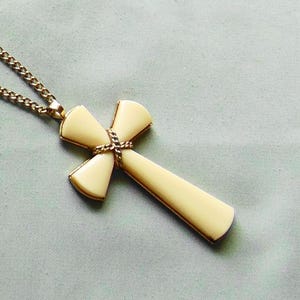 May include: A cross pendant necklace featuring a cream-colored, smooth-surfaced cross with flared ends. The center of the cross is wrapped with a twisted gold-tone metal detail, and it hangs from a matching gold-tone metal link chain.