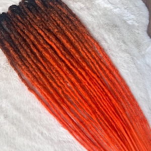 May include: A set of synthetic hair extensions featuring a dark brown to vibrant orange ombre color gradient. The individual strands are styled into thin, textured dreadlocks that transition from a deep dark brown at the top to bright orange.