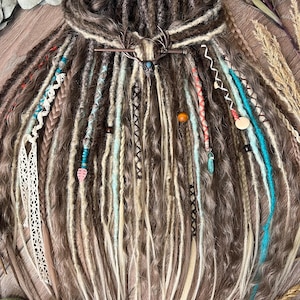 Op de afbeelding: Een set bruine en blonde dreadlocks met turquoise, roze en crèmekleurige accenten. De dreadlocks zijn versierd met kralen, vlechten en een decoratief metalen haaraccessoire. Het haar is gestyled met een mix van texturen en kleuren.
