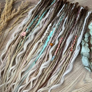 Może przedstawiać: Zestaw syntetycznych dredów w odcieniach brązu, blondu i bieli z niebieskimi i brązowymi akcentami. Dredy są ozdobione koralikami, kryształami i koronką.