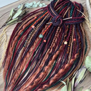 May include: A collection of colorful dreadlocks in shades of burgundy, brown, and peach, accented with green and orange strands. The dreads are styled with decorative beads and a wooden hair pin, creating a bohemian aesthetic.