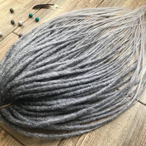 May include: A set of synthetic dreadlocks featuring a color gradient that transitions from a dark charcoal gray at the base to a light silver gray at the tips. The dreadlocks are bundled together with a band and include several small beads.