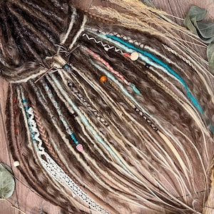 May include: A close-up of brown, blonde, and teal dreadlocks with decorative beads and wraps. The dreadlocks are styled with a metal hair accessory and various embellishments. The overall aesthetic is bohemian and natural.