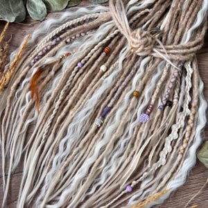 May include: A set of synthetic dreadlocks in shades of beige, tan, and white, featuring wavy and straight textures. The bundle includes braided sections, wrapped accents, and decorative beads in purple, brown, and cream, with a small turtle charm.