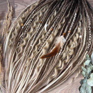 Könnte beinhalten: Ein Set aus synthetischen Dreadlocks in Blond- und Brauntönen mit welligen und glatten Texturen. Die Haarverlängerungen sind mit weißen Garnwicklungen, Holzperlen, einem hornförmigen Anhänger und einer braun-weißen Feder verziert.