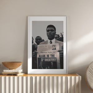 Puede incluir: Impresión en blanco y negro enmarcada que presenta a Muhammad Ali sosteniendo un periódico con el titular "ALLAH IS THE GREATEST". Ali lleva un traje y corbata, con otras personas en el fondo. La impresión se exhibe en un mueble blanco.