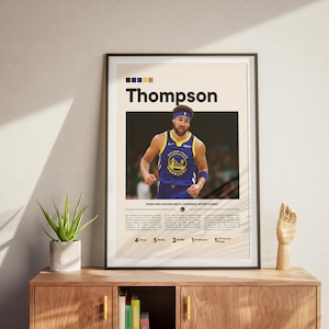 May include: Framed poster featuring basketball player Thompson in a blue and gold jersey. The poster includes the player's name, stats, and team logo. The poster is displayed on a wooden cabinet with a plant and a wooden hand sculpture.
