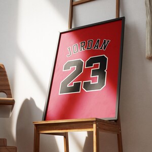以下が含まれることがあります： 赤色のフレームプリント。黒と白で「JORDAN」の文字と「23」の数字が描かれています。木製の椅子に飾られており、壁に立てかけられています。ミニマリストでモダンなデザインで、インテリアに適しています。
