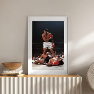 Puede incluir: Fotografía enmarcada de un combate de boxeo. Un boxeador con pantalones cortos blancos y guantes rojos se encuentra sobre un oponente caído con pantalones cortos y guantes negros. La imagen está ambientada en un ring de boxeo con público al fondo.