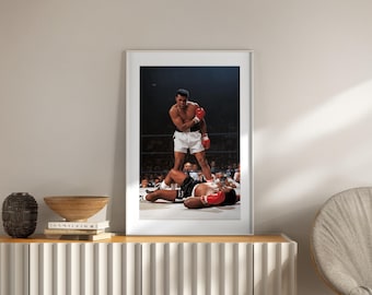 Puede incluir: Fotografía enmarcada de un combate de boxeo. Un boxeador con pantalones cortos blancos y guantes rojos se encuentra sobre un oponente caído con pantalones cortos y guantes negros. La imagen está ambientada en un ring de boxeo con público al fondo.