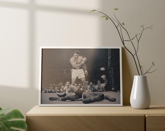Puede incluir: Fotografía enmarcada en blanco y negro de un combate de boxeo. Un boxeador está de pie sobre otro que está tendido en la lona. La fotografía se exhibe sobre una superficie de madera con un jarrón blanco y una planta.