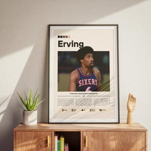 May include: Framed poster featuring basketball player Erving in a Sixers jersey. The poster has a cream background with black text and a photo of the player. The poster is displayed on a wooden cabinet with a plant and a wooden hand sculpture.