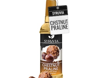 May include: A clear glass bottle of Syruvia Chestnut Praline pure cane syrup with a gold cap. The label features images of chestnuts and a praline candy. A rectangular white tag attached to the neck displays the text SYRUVIA CHSTNUT PRALINE.