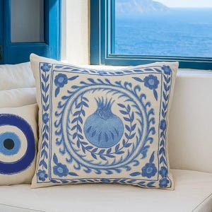 May include: Square decorative pillow with a cream background and a blue pomegranate design. The pomegranate is surrounded by a wreath of leaves and flowers. Another pillow with a blue eye design is visible.
