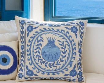 Puede incluir: Cojín cuadrado crema con un diseño de granada azul rodeado de una corona de hojas y flores. Se ve otro cojín con un motivo de ojo azul. La escena está ambientada cerca de una ventana con vistas al mar.