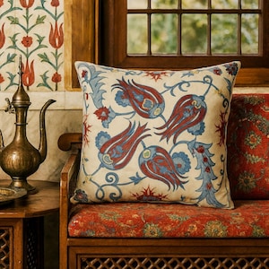May include: A square decorative pillow with a cream background and a repeating pattern of red and blue tulip-like flowers. The pillow is placed on a wooden bench with a red patterned cushion. The scene is set near a window.