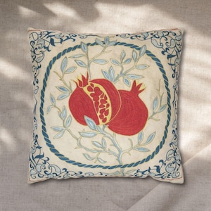 May include: Square decorative pillow with a cream-colored background. The pillow features a central design of two red pomegranates with yellow accents and blue leaves. A blue rope-like border and decorative blue corner patterns complete the design.