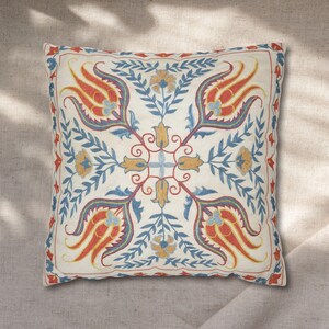 May include: Square decorative pillow with an embroidered floral pattern. The design features stylized red, yellow, and blue flowers and leaves on a cream-colored background. The pillow has a red border.