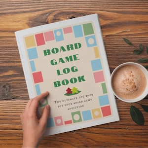 以下が含まれることがあります： カラフルな幾何学模様と「BOARD GAME LOG BOOK」の文字が入ったクリーム色の表紙のボードゲームログブック。本は開かれており、手が見え、木製の表面にコーヒーカップが置かれています。