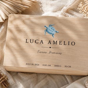 May include: A rectangular light-colored wooden keepsake box featuring a watercolor blue sea turtle illustration. The text reads LUCA AMELIO, Forever Memories, JULY 18, 2026, 6:00 AM, 3000 G, and 50 CM, with a small heart icon centered below the name.