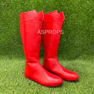 Może przedstawiać: Para jaskrawoczerwonych butów do kolan. Buty wykonane są z gładkiego materiału i mają płaską podeszwę. Prezentowane są na zielonej powierzchni. Na butach widoczny jest napis "ASPROPS".