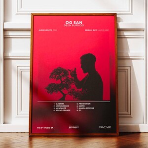 Peut inclure: Affiche encadrée avec un fond rouge et le texte "OG SAN DEEN BURBIGO". Une silhouette noire d'une personne taillant un bonsaï est au centre. L'affiche comprend une liste de titres et une date de sortie. Le cadre est marron.