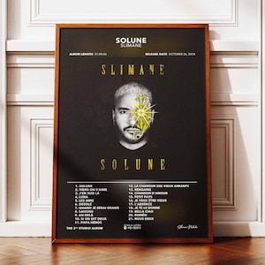 Puede incluir: Un póster enmarcado del álbum "Solune" de Slimane. El póster presenta un retrato del artista con un diseño dorado en su rostro. El título del álbum y la lista de canciones se muestran en texto dorado sobre un fondo negro.