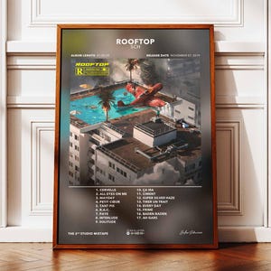 Op de afbeelding: Een ingelijste poster voor het album "ROOFTOP" van SCH. De illustratie toont een surrealistisch stadsbeeld met een rood vliegtuig en een zwembad. De poster bevat de tracklist van het album en de releasedatum.