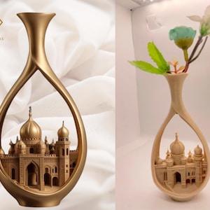 May include: Gold-colored decorative vase with an open, teardrop-shaped design. The vase features a detailed relief of a building with domes and arches. The vase on the right holds artificial flowers. The Ayat Artworks logo is in the upper left corner.