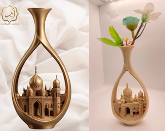 May include: Gold-colored decorative vase with an open, teardrop-shaped design. The vase features a detailed relief of a building with domes and arches. The vase on the right holds artificial flowers. The Ayat Artworks logo is in the upper left corner.