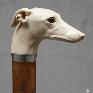 May include: A walking cane with a sculpted ivory-colored dog head handle. The dog has brown eyes and a long snout. The handle is attached to a brown wooden shaft with a silver band that has the letters "SON." engraved on it.