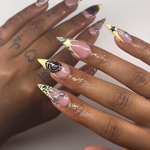 Peut inclure: Longs ongles pointus avec une base rose clair, des pointes jaunes et des motifs argentés et noirs. Les motifs comprennent des spirales, des étoiles et des formes abstraites. Les ongles sont bien manucurés et semblent être en acrylique ou en gel.