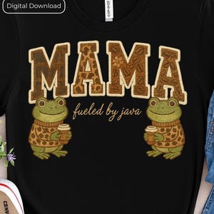 May include: Black t-shirt with the word "MAMA" in a brown, coffee bean-patterned font. Two green frog illustrations wearing sweaters and holding coffee cups flank the text. The phrase "fueled by java" is below the word "MAMA".