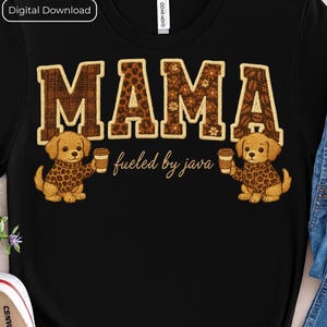 May include: Black t-shirt with the word "MAMA" in brown, coffee bean-filled letters. Two cartoon puppies wearing leopard print sweaters and holding coffee cups flank the text. The phrase "fueled by java" is below the word "MAMA".