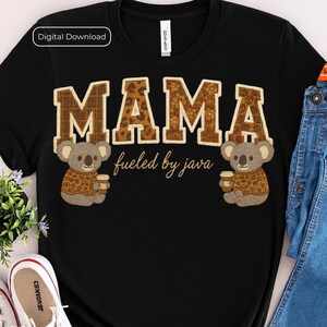 May include: Black t-shirt with the word "MAMA" in a brown, textured font with leopard print accents. Two cartoon koalas wearing sweaters and holding coffee cups flank the text. The phrase "fueled by java" is written below.