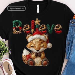 Może przedstawiać: Czarny t-shirt ze słowem "Believe" w zielonych, lamparcich i czerwonych literach. Uroczy lis w czapce Świętego Mikołaja jest umieszczony poniżej. Obraz ma etykietę "Pobieranie cyfrowe".