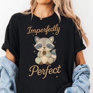 Könnte beinhalten: Schwarzes T-Shirt mit einem Cartoon-Waschbären-Design. Der Waschbär ist mit geschlossenen Augen und gefalteten Händen dargestellt, hergestellt aus Patchwork-Stoff in Grau, Blau und Beige. Die Worte "Imperfectly Perfect" sind in Gold über und unter dem Waschbären geschrieben.