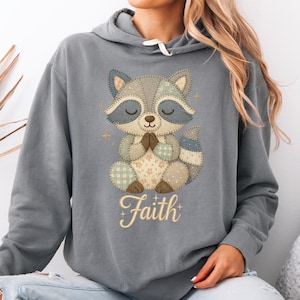 Könnte beinhalten: Grauer Kapuzenpullover mit einem Cartoon-Waschbären-Design. Der Waschbär betet, mit einem Patchwork-Design und dem Wort "Faith" in Schrift. Der Waschbär hat blaue und graue Akzente mit goldenen Sterndetails.