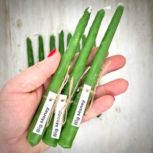 May include: A set of slender green taper candles, each featuring a white paper tag tied with twine that reads Big Money. The candles have a smooth, cylindrical shape with a pointed top and a white wick, presented in a group of several items.