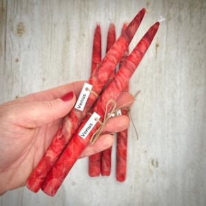 May include: Red and white marbled taper candles. Each candle has a tag that reads "Venus" and is tied with twine. The candles are displayed on a light wood surface.