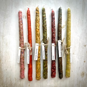 May include: An overhead shot of eight colorful, textured taper candles. Each candle is a different color, ranging from pink to green to brown. Small white tags with text like "Intuition" and "Personal Power" are tied to each candle.