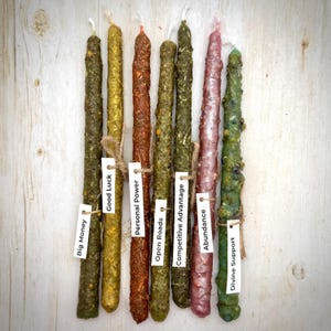 May include: A collection of nine colorful, handmade spell candles. Each candle is a different color, including green, gold, and red, and has a small tag with a word like "Good Luck" or "Abundance". The candles are arranged on a light wood surface.