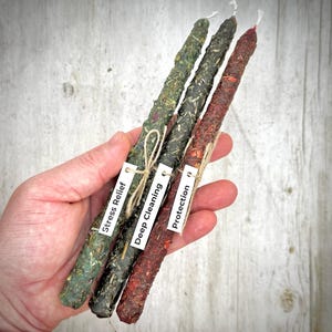 May include: Three slender taper candles coated in dried herbs and botanicals. The candles are green, dark charcoal, and deep red. Each candle features a small white paper tag tied with twine, labeled Stress Relief, Deep Cleaning, and Protection.