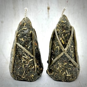 May include: Two dark green, cone-shaped candles with a textured surface and a light-colored, crisscross design. The candles are filled with straw-like material and have a white wick at the top. The background is a light, textured surface.