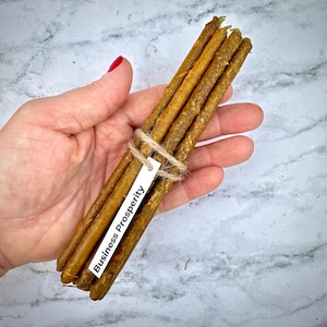 Può includere: Un fascio di candele sottili marroni legate con spago. Un'etichetta bianca con le parole "Business Prosperity" è attaccata. Le candele hanno un aspetto strutturato e rustico, possibilmente realizzate in cera naturale.