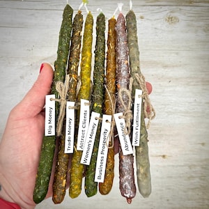 May include: A collection of eight spell candles in various colors, including green, yellow, and brown. Each candle is adorned with herbs and tied with twine, and labeled with phrases like "Big Money" and "Wish Fulfillment".